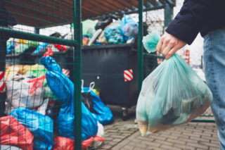 Hand with garbage against full trash cans with rubbish bags overflowing onto the pavement.